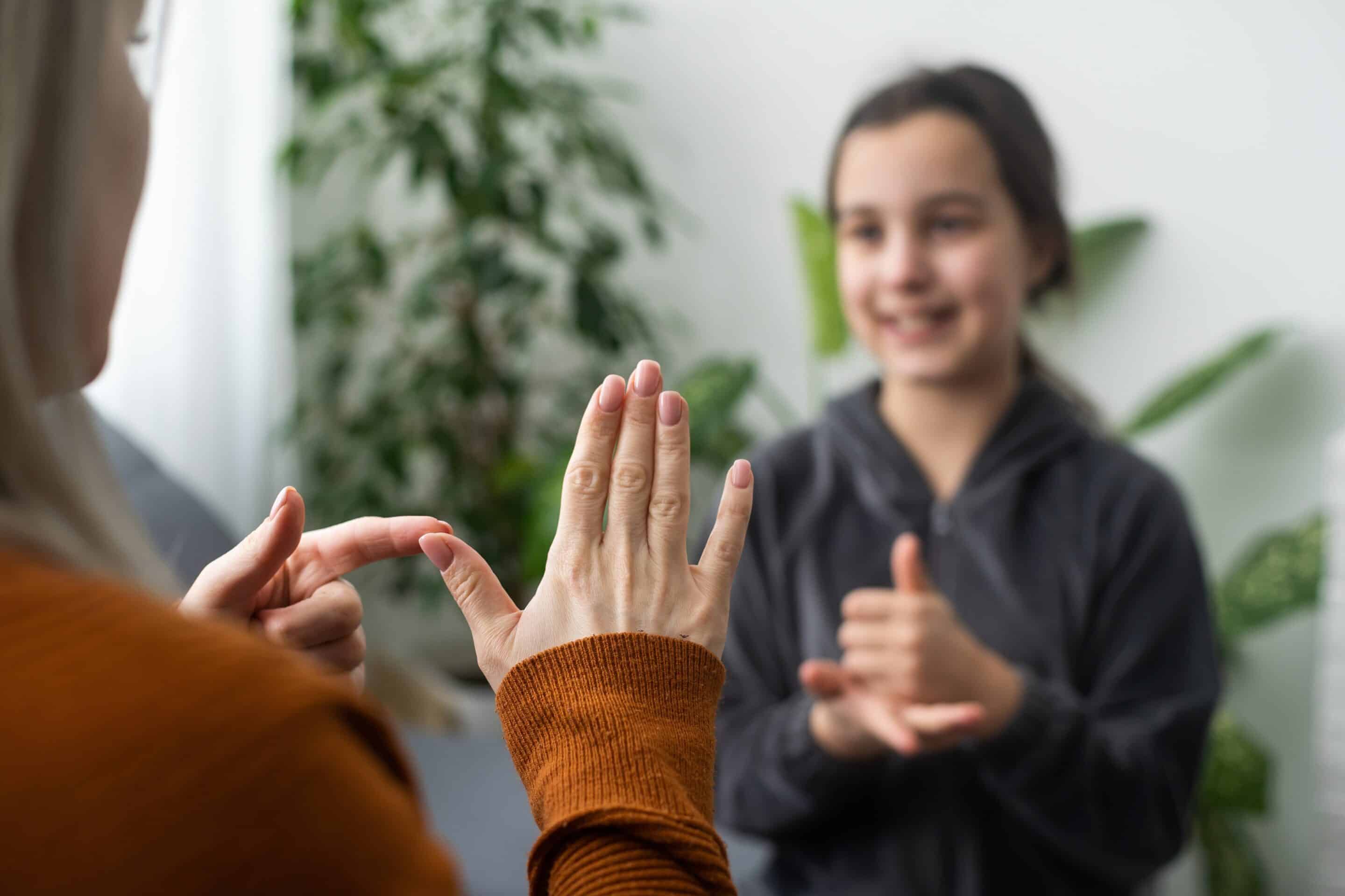 Criança surda usando LIBRAS, gesticulando ao lado de terapeuta com tablet exibindo sinais